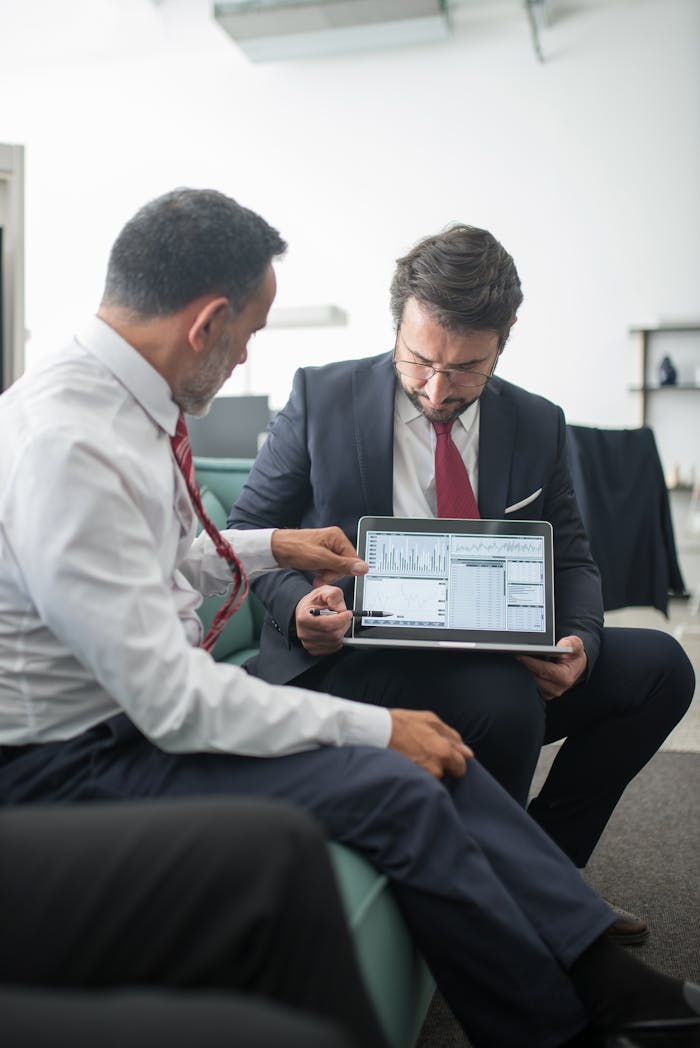 Two businessmen in an office reviewing economic data on a laptop screen during a meeting.